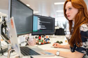 Red haired woman with freckles sitting at a desk with a laptop and two external monitors. She is writing code inside VS Code program. She is looking at her laptop screen which is on her left.