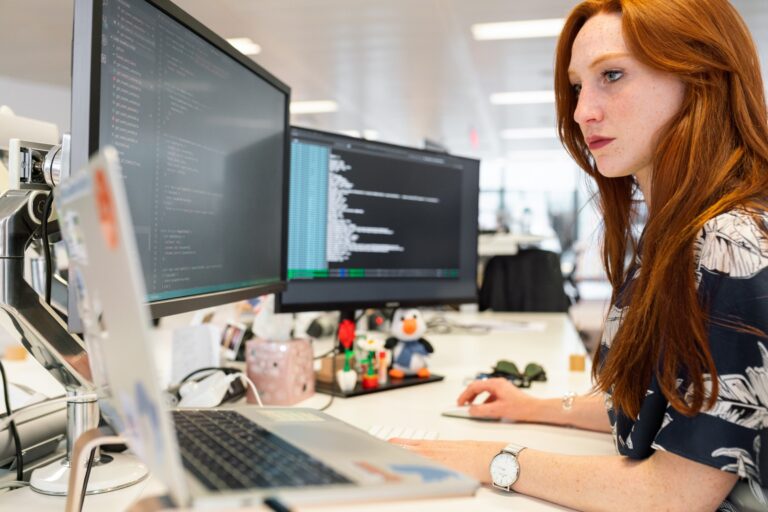 Red haired woman with freckles sitting at a desk with a laptop and two external monitors. She is writing code inside VS Code program. She is looking at her laptop screen which is on her left.