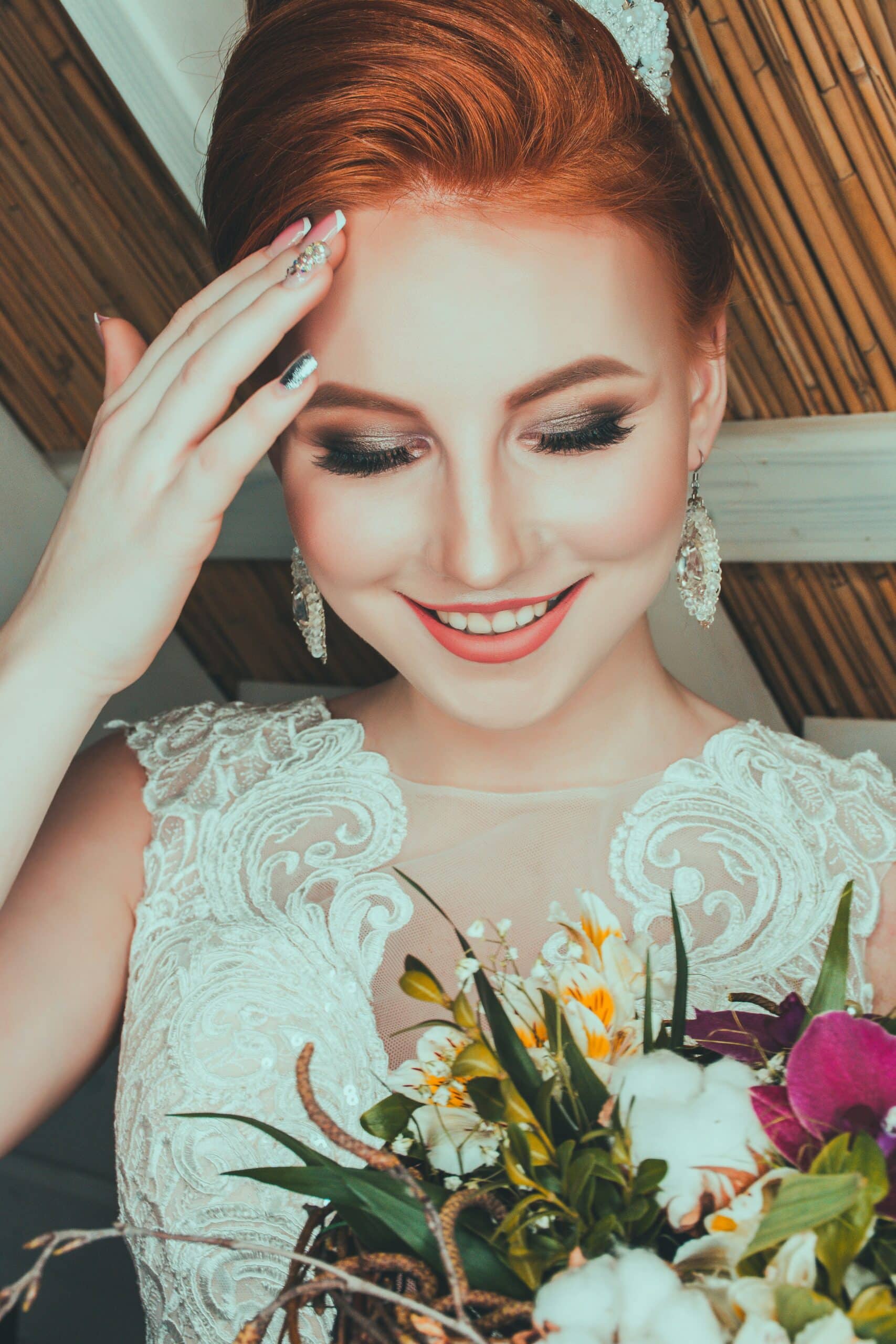 The image consists of a redheaded woman, smiling. Her right-hand with beautiful nails is touching her forehead. She is wearing white diamond earings. She is wearing a cap sleeved white wedding dress with floral designs. In her left hand she is holding a floral bouquet slightly off-screen.